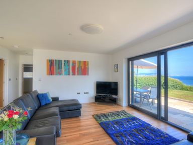 A living room with a black sectional sofa a colorful rug and a television near large sliding glass doors overlooking a patio and ocean at Harbour Heights Cottage in Lyme Regis