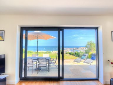 A room with glass sliding doors leading to a patio with table chairs umbrella and sun loungers overlooking the sea at Harbour Heights Cottage in Lyme Regis