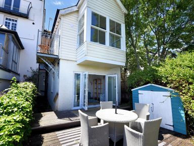 An outdoor patio area with a round table and four chairs next to a blue and white shed and a two-story house with white siding and large windows at Little Bay View in Lyme Regis
