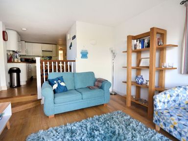 A living room with blue sofa, patterned armchair, wooden shelving unit, and kitchen area at Little Bay View in Lyme Regis