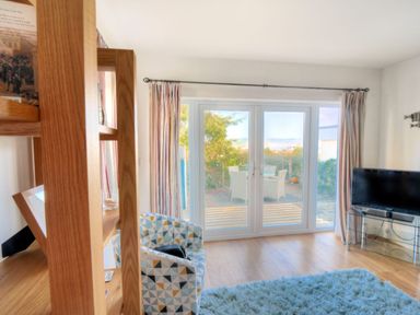 A living room with a patterned armchair a wooden shelf a television on a glass stand and glass doors leading to an outdoor patio at Little Bay View in Lyme Regis
