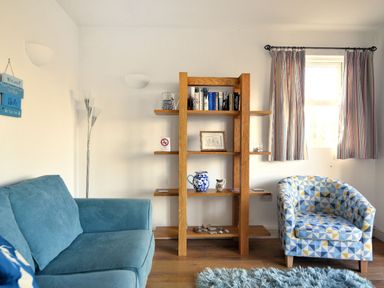 A living room with a blue sofa a patterned armchair a wooden bookshelf and a window with striped curtains at Little Bay View in Lyme Regis