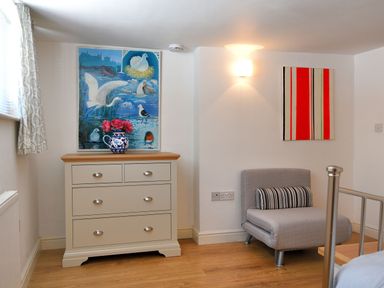 A room with a chest of drawers topped with a vase of flowers an armchair with a striped cushion and paintings on the walls at Little Bay View in Lyme Regis