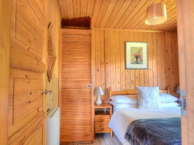 A bedroom with a wooden bed white bedding a bedside table with a lamp wooden walls and a framed picture at 4 Pinewood Retreat in Pinewood