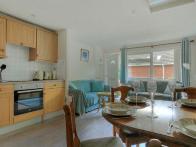 A kitchen with wooden cabinets and a dining table set with dishes and glasses next to a living area with blue sofas and large windows at 36 Fernhill Heights in Charmouth