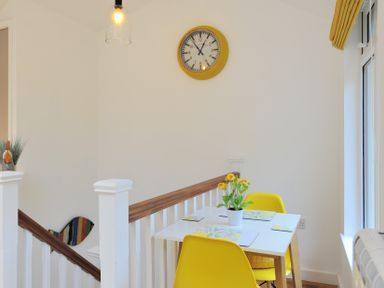 A dining area with a white table yellow chairs yellow flowers in a pot a wall clock and a staircase at Seashore Lyme Regis