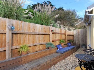 An outdoor seating area with wooden bench cushions potted plants a wooden fence and a small round metal table at Seashore in Lyme Regis