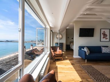 A room with large windows overlooking the sea a blue sofa a brown armchair a chest of drawers and a wall-mounted tv at 3 Beach Way House in Lyme Regis