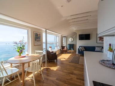A room with a dining table and chairs near large windows overlooking the sea with armchairs a sofa a TV and a kitchen counter at 3 Beach Way House in Lyme Regis