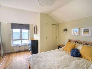 A bedroom with a bed with yellow and blue pillows a chest of drawers a window with a view of the sea and framed pictures on the wall at 3 Beach Way House in Lyme Regis