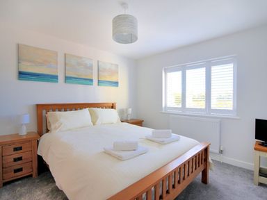 A bedroom with a wooden bed dressed in white linens two bedside tables with lamps three paintings above the bed a window with white shutters and a small TV on a stand at Shire View in Lyme Regis