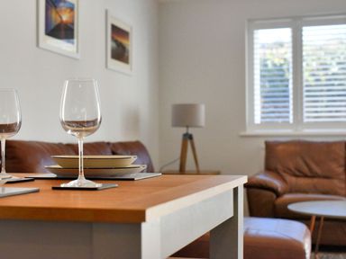 A dining table with wine glasses and plates in a living room with brown leather furniture and a window with blinds at Shire View in Lyme Regis