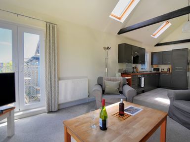 A living room with armchairs a wooden coffee table with bottles a television and a kitchen with dark cabinets at Bay View in Lyme Regis
