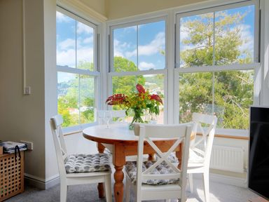 A dining area with a round wooden table and white chairs near large windows with a view of trees at Bay View in Lyme Regis