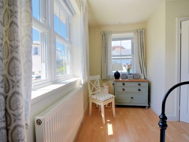 A small room with two windows a white chair a chest of drawers with a lamp and flowers on it and a radiator under one window at Bay View in Lyme Regis