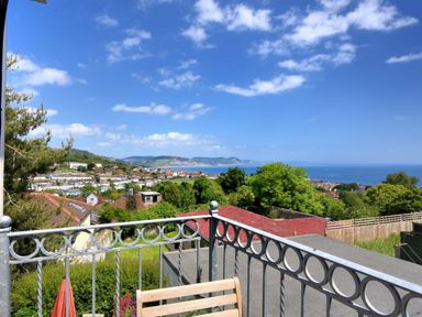 A balcony with a metal railing and wooden chair overlooking houses trees hills and the sea at Bay View in Lyme Regis