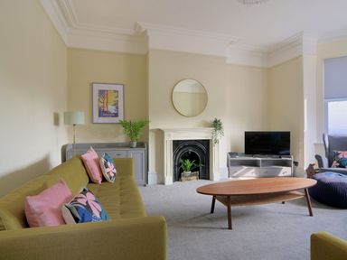 A living room with a green sofa pink and floral cushions a wooden coffee table a fireplace with plants and a round mirror at 7 Seafield Road in Seaton