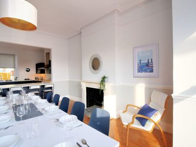 A dining room with a white table set with bowls and glasses and navy chairs next to a kitchen with bar stools and a white armchair with a blue cushion at 7 Seafield Road in Seaton