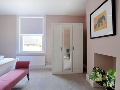A bedroom with a wardrobe, a pink bench, a window with a roller blind, a framed elephant print, and a potted plant at 7 Seafield Road Seaton