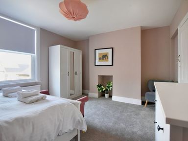 A bedroom with a white bed with towels on it a white wardrobe a framed picture above a small alcove with a plant a gray chair and a white dresser at 7 Seafield Road in Seaton