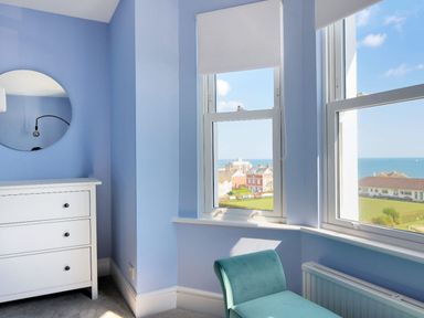 A corner of a room with a white chest of drawers a round wall mirror two windows with a sea view and a teal chair at 7 Seafield Road Seaton
