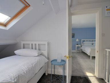 A small bedroom with a single bed under a skylight and a blue side table next to an open door leading to a larger bedroom with a double bed and bedside table at 7 Seafield Road Seaton