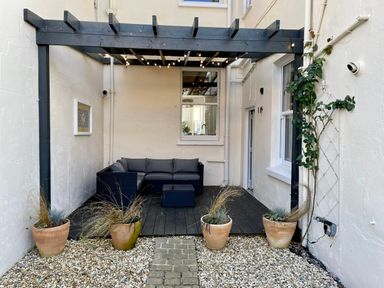 An outdoor patio area with a black pergola string lights a black sectional sofa a small table four potted plants gravel and a stone path at 7 Seafield Road in Seaton