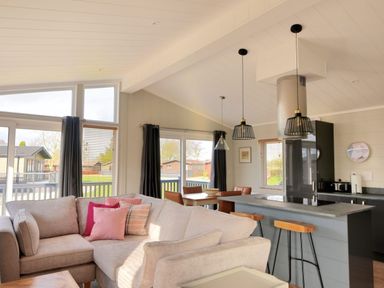 An open living room and kitchen area with a beige sectional sofa with pink cushions a kitchen island with stools pendant lights and large windows at Hazelwood 4 @ Pinewood Retreat in Pinewood