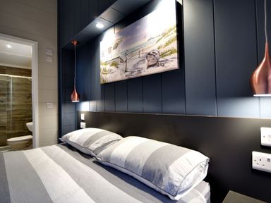A bedroom with a bed with striped bedding a painting of a beach scene above the headboard and an open door showing a bathroom at Hazelwood 4 @ Pinewood Retreat in Pinewood