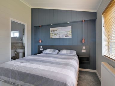A bedroom with a double bed with striped bedding a blue paneled wall with a painting two hanging pendant lights a window with blinds and an en suite bathroom at Hazelwood 4 @ Pinewood Retreat Pinewood