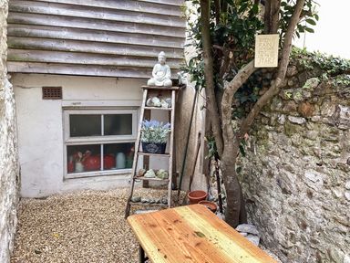 A small outdoor space with gravel ground a wooden table a wooden shelf with a Buddha statue plants and stones a tree with a small sign on a stone wall at The Little Rose in Lyme Regis