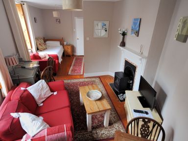 A living room with a red sofa white pillows a wooden coffee table a rug a fireplace a TV and a single bed in the background at Driftwood in Lyme Regis