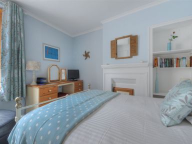 A bedroom with a bed a desk with a lamp a small television and built in shelves with books and vases at Driftwood in Lyme Regis