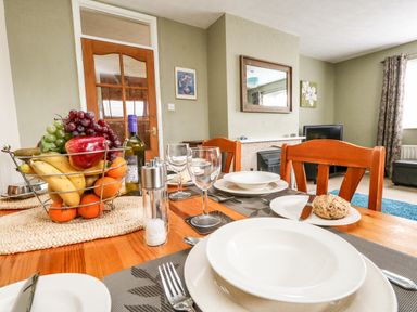 A dining table with plates glasses and cutlery next to a fruit basket in a living room with a fireplace mirror and television at Sunny Haven in Preston