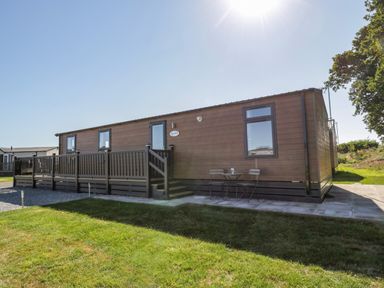 A single story brown wooden mobile home with a fenced porch and a small outdoor table with two chairs at Away Daze in Carnaby near Bridlington