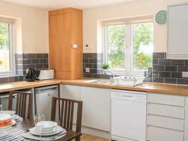 A kitchen with white cabinets wooden countertops a dishwasher a toaster a kettle and a dining table with plates and glasses at Sawtons Cottage 2 in Dawlish