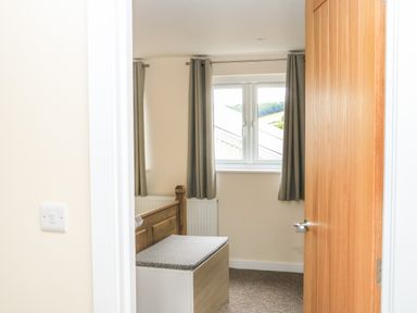 A bedroom with a wooden bed frame a bench with a cushion a window with curtains and a door at Sawtons Cottage 2 in Dawlish