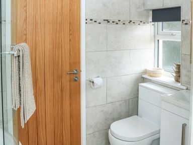 A bathroom with a wooden door white toilet gray tiled walls and a window with a roller blind at Sawtons Cottage 2 in Dawlish