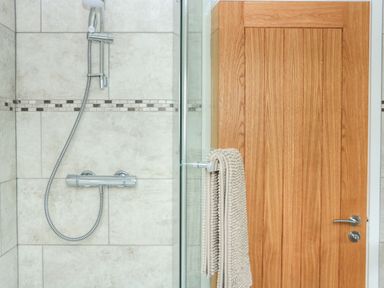 A tiled shower area with a silver showerhead and faucet next to a wooden door with a beige towel hanging on the handle at Sawtons Cottage 2 in Dawlish