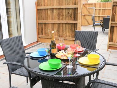 An outdoor patio dining area with a glass table set with colorful bowls and plates wine glasses and a bottle of champagne at Sawtons Cottage 2 in Dawlish