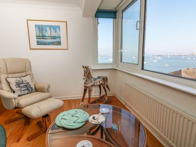 A room with a beige recliner chair and footrest a wooden rocking horse a glass round coffee table and a window showing a sea view at Port View in Falmouth