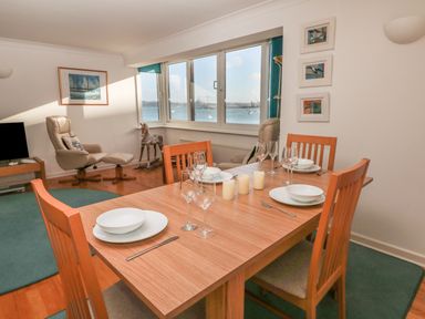 A dining area with a wooden table set with white plates and glasses next to a sitting area with beige chairs and a view of the sea at Port View in Falmouth