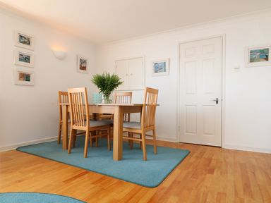 A dining area with a wooden table and four chairs on a blue rug with framed pictures on the walls at Port View in Falmouth