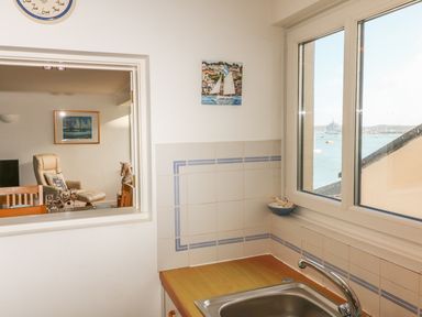 A kitchen sink under a window with a view of the sea and a room with chairs and a painting beyond the kitchen window at Port View in Falmouth