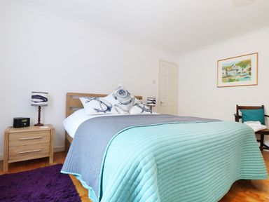 A bedroom with a bed covered by blue and gray blankets a wooden nightstand with a lamp and clock a wooden chair with a cushion and a framed painting on the wall at Port View in Falmouth