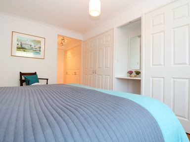A bedroom with a bed covered by a blue bedspread a chair with a blue cushion a wall painting built-in wardrobes and a dressing table at Port View in Falmouth