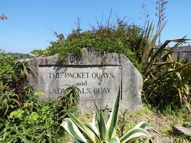 A stone sign with the text The Packet Quays and Admirals Quay surrounded by plants