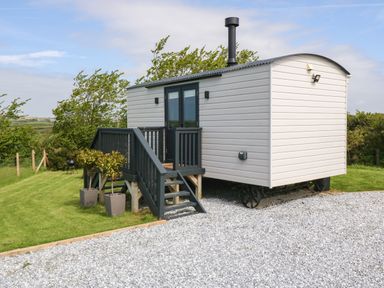 A small white wagon with a black chimney and black stairs on a gravel driveway surrounded by grass and bushes at Cwt Y Rhos in Valley