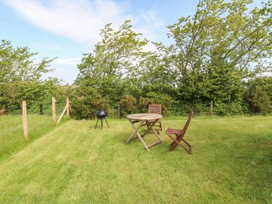 A grassy backyard with a round wooden table, two wooden chairs, a small black grill and a wire fence with trees in the background at Cwt Y Rhos Valley