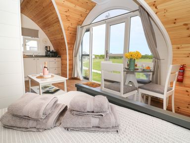 An interior view of a wooden pod with a bed, folded towels, a small kitchen area, a table with chairs, and a glass door leading outside at Poppy Seaton near Hornsea